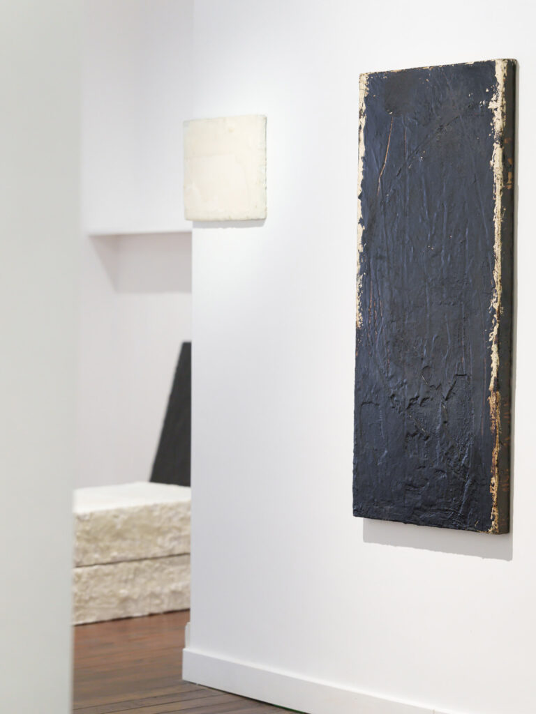 Installation View of Eleanor Bartlett at Vanner Gallery, Painting on the right: Scored I, 2000, Tar and metal paint on canvas, 122 x 61cm, Painting left on the wall: Small painting, 2021, Wax on canvas, 30 x 41cm, Foreground on the floor: Wax tablets I & II, 2023 , Wax on wood and canvas, 20 x 77 x 61cm (each), Background on the floor: Black painting, 2023, Tar and metal paint on canvas, 90 x 61cm © Image Courtesy the gallery, Photography by Ash Mills